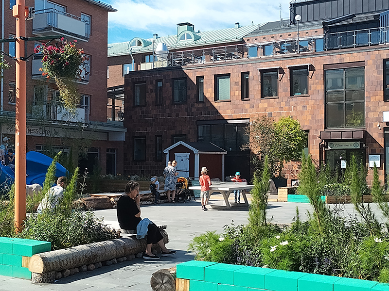 Bild över torget under sommaren 2024. Ett pingisbord står i mitten där två barn spelar. En kvinna sitter i förgrunden på en upphöjd plantering
