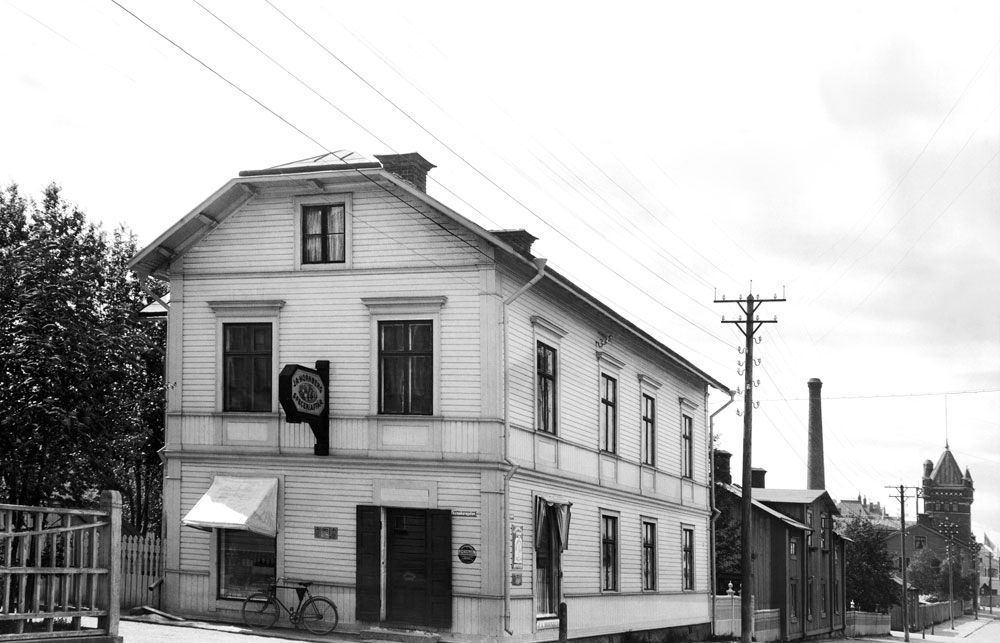 Vit träbyggnad i två våningar. Butikslokal på gaveln.