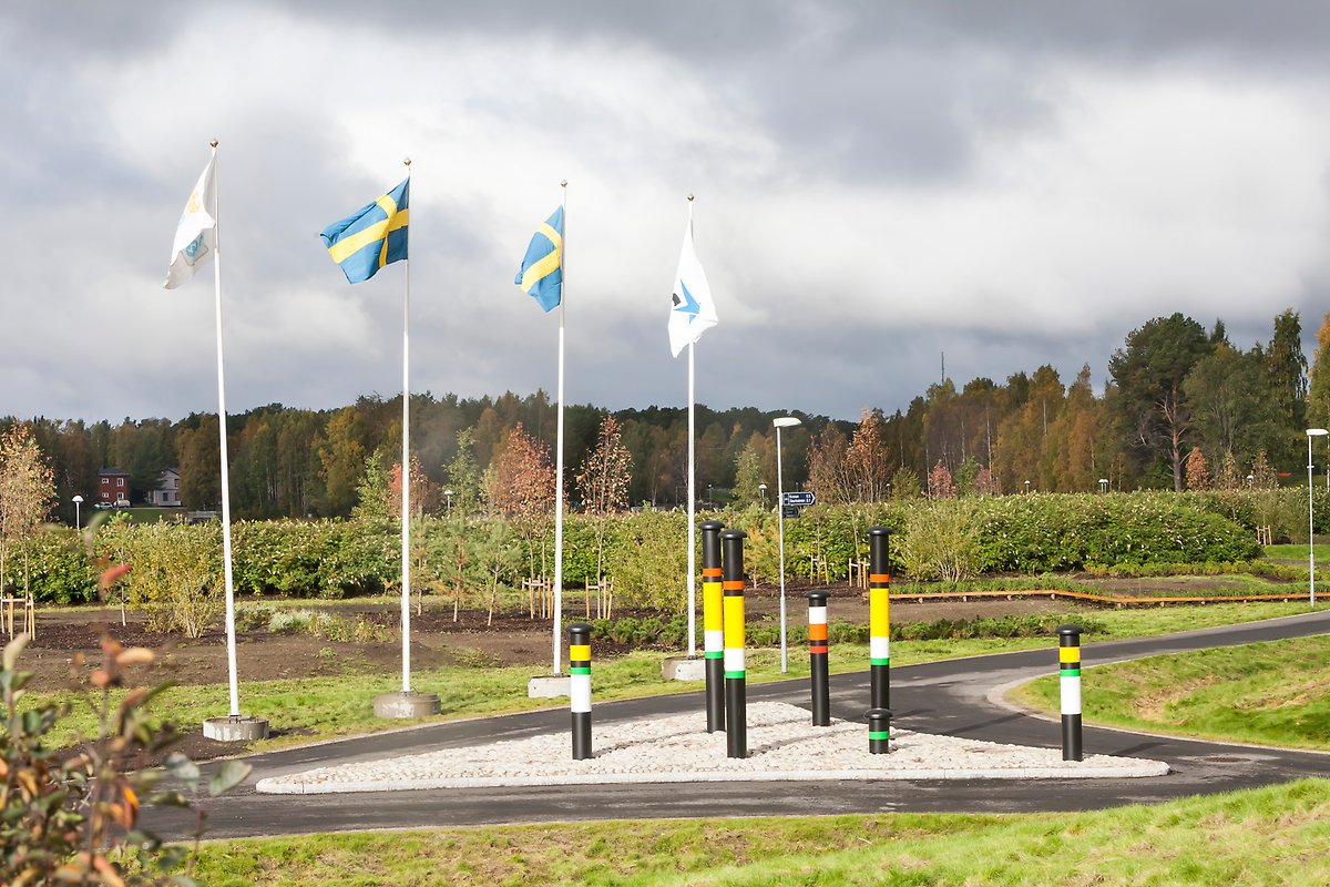 Flaggstänger  med svenska flaggan hissade. Framför dem syns en trekantig sandyta med ventilationsrör från vatten och avloppsanläggningar. De svarta rören är målad i vitt, grönt, gult och rött.